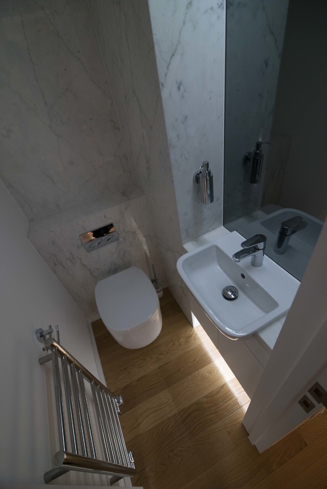 This is an example of a small contemporary cloakroom in London with flat-panel cabinets, white cabinets, a wall mounted toilet, stone slabs, light hardwood flooring, a built-in sink, marble worktops and white tiles.