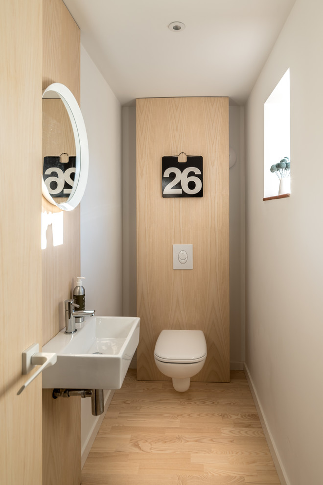 Nordisk inredning av ett vit vitt toalett, med en vägghängd toalettstol, vita väggar, mellanmörkt trägolv, ett väggmonterat handfat och brunt golv