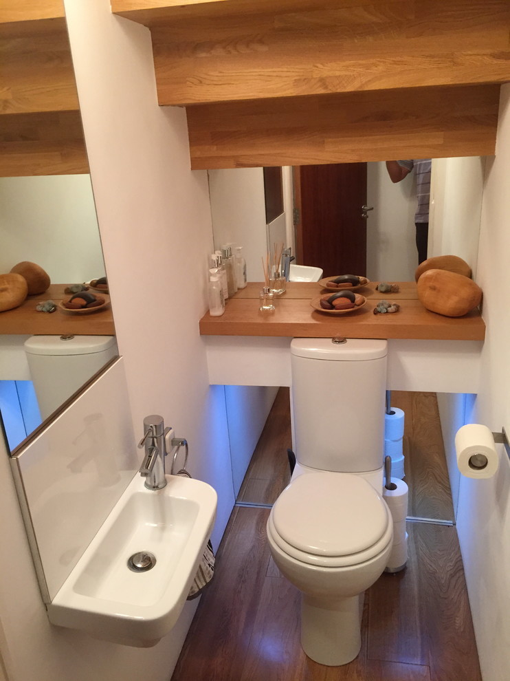 Inredning av ett stort toalett, med ett väggmonterat handfat, träbänkskiva, en toalettstol med hel cisternkåpa, vita väggar och mellanmörkt trägolv