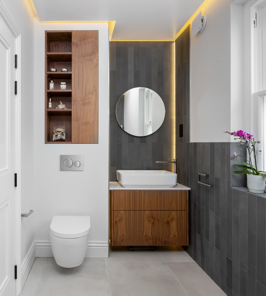 Inredning av ett skandinaviskt vit vitt toalett, med släta luckor, skåp i mellenmörkt trä, en vägghängd toalettstol, grå kakel, vita väggar, ett fristående handfat och grått golv