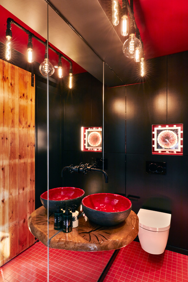 Bild på ett litet funkis brun brunt toalett, med släta luckor, svarta skåp, en vägghängd toalettstol, svart kakel, svarta väggar, klinkergolv i porslin, ett fristående handfat, träbänkskiva och rött golv