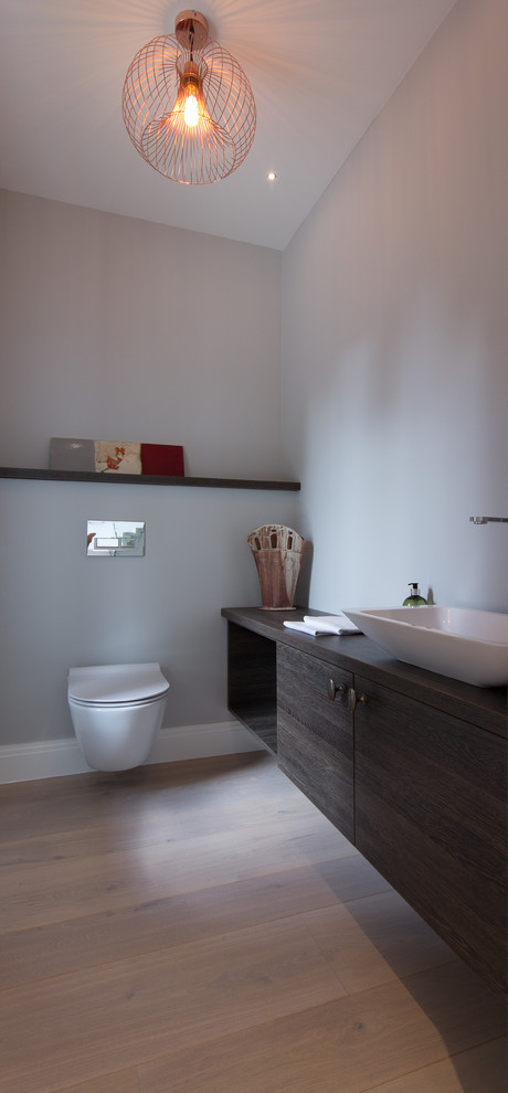 Photo of a medium sized modern cloakroom in London with flat-panel cabinets, dark wood cabinets, a wall mounted toilet, grey walls, medium hardwood flooring, a console sink, wooden worktops and brown floors.