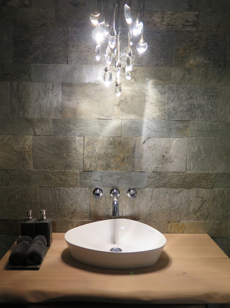 Inredning av ett modernt litet toalett, med en vägghängd toalettstol, grå kakel, stenkakel, grå väggar, ett väggmonterat handfat och träbänkskiva
