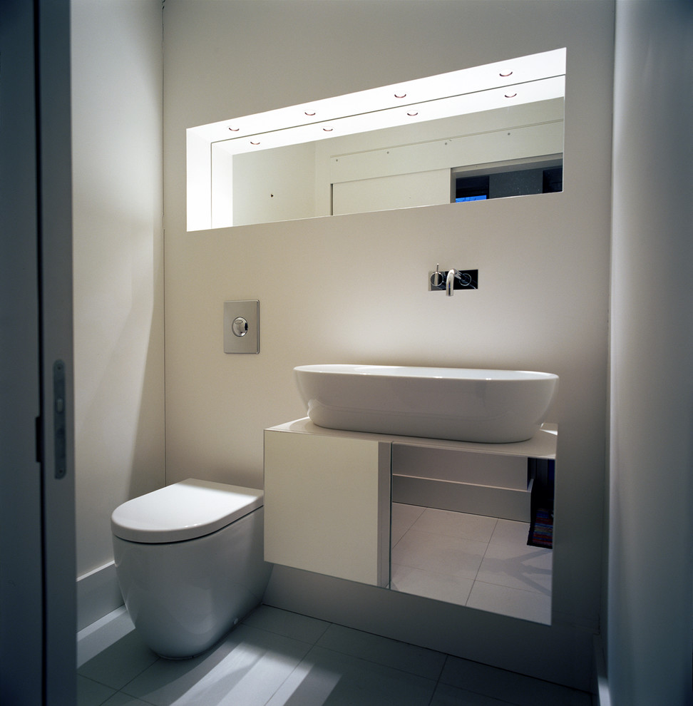 Idée de décoration pour un grand WC suspendu design avec un placard à porte plane, des portes de placard blanches, des carreaux de miroir, un mur blanc, un sol en carrelage de porcelaine, une vasque, un sol blanc et un plan de toilette blanc.