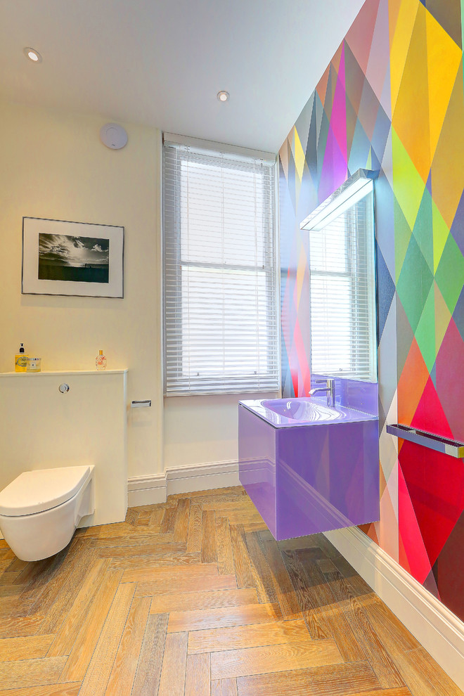 This is an example of a large contemporary cloakroom in London with a wall mounted toilet, multi-coloured walls, light hardwood flooring, a wall-mounted sink, glass worktops, beige floors and purple worktops.