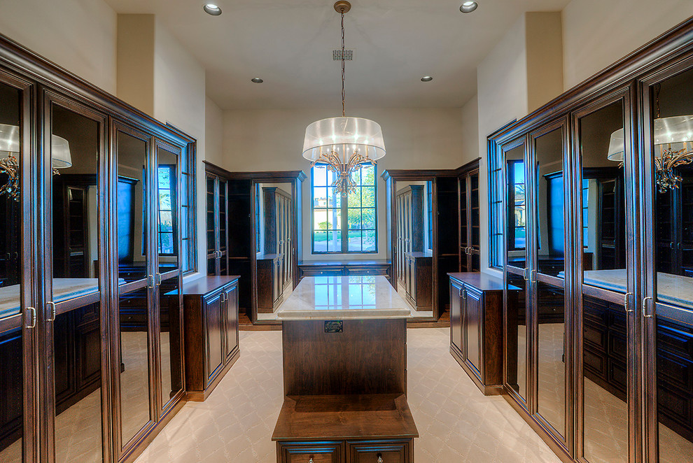 Photo of an expansive mediterranean gender neutral walk-in wardrobe in Phoenix with glass-front cabinets, dark wood cabinets, carpet and beige floors.