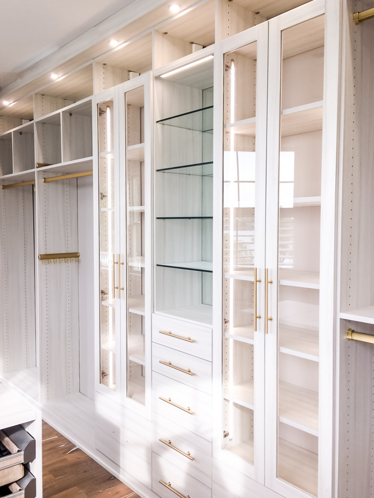 На фото: большая гардеробная комната в стиле неоклассика (современная классика) с стеклянными фасадами, белыми фасадами, паркетным полом среднего тона и коричневым полом
