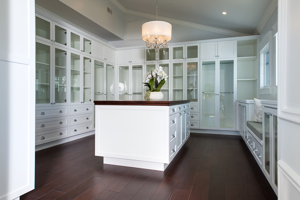 This is an example of an expansive beach style gender neutral walk-in wardrobe in Los Angeles with glass-front cabinets, white cabinets and dark hardwood flooring.