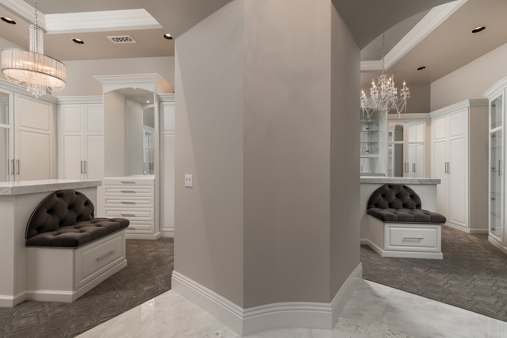 This is an example of an expansive mediterranean gender neutral walk-in wardrobe in Phoenix with glass-front cabinets, white cabinets, carpet, grey floors and a coffered ceiling.