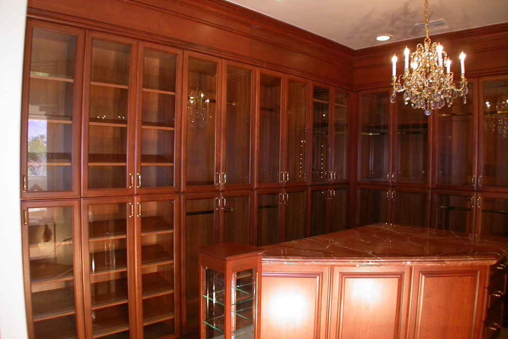 Large classic gender neutral walk-in wardrobe in Las Vegas with glass-front cabinets, dark wood cabinets and carpet.