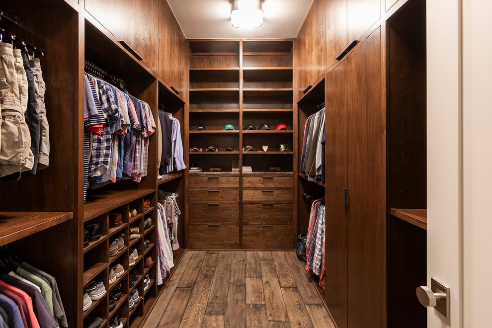Modelo de armario vestidor de hombre actual de tamaño medio con armarios con paneles lisos, puertas de armario de madera oscura y suelo de madera en tonos medios
