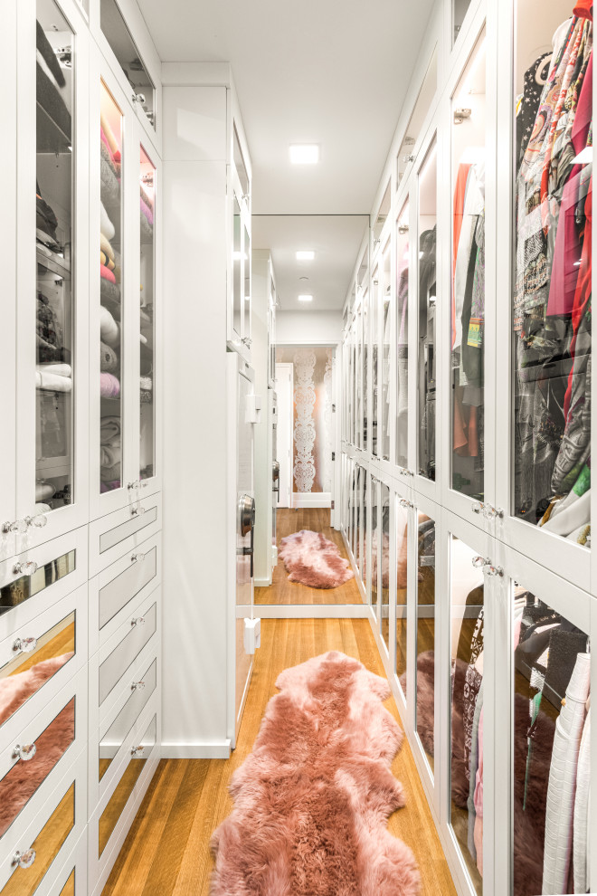 Modern inredning av ett stort walk-in-closet för kvinnor, med luckor med glaspanel, vita skåp och mellanmörkt trägolv