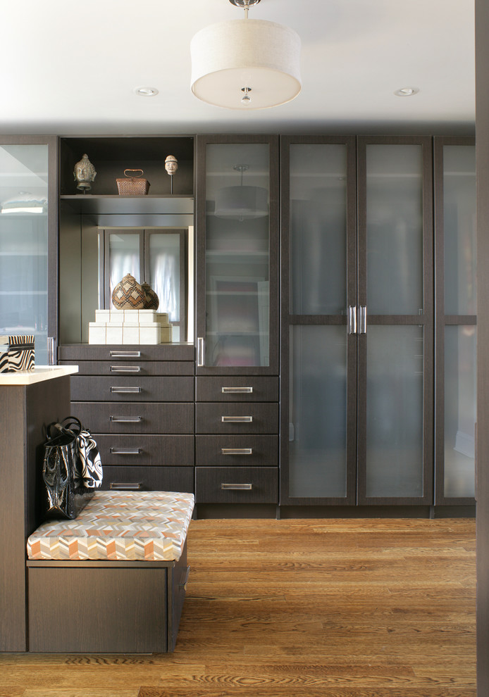 Cette image montre un dressing room design en bois foncé neutre avec un placard à porte vitrée et un sol en bois brun.