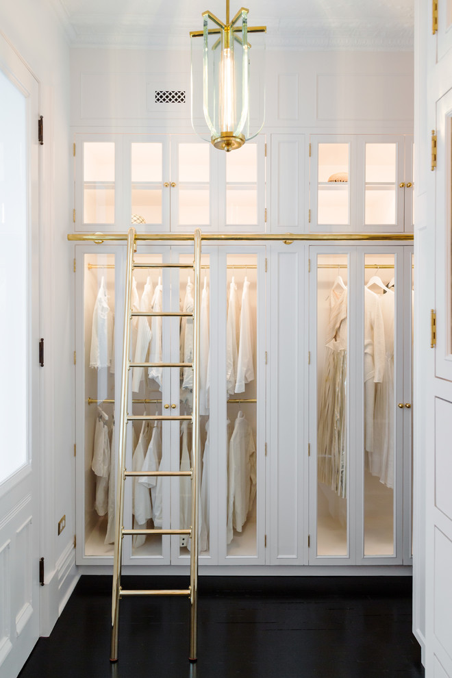 Victorian walk-in wardrobe in New York with glass-front cabinets, white cabinets and dark hardwood flooring.