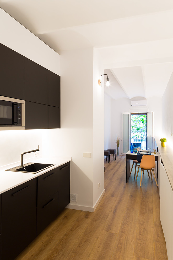 Example of a small danish single-wall light wood floor and brown floor open concept kitchen design in Barcelona with a single-bowl sink, flat-panel cabinets, black cabinets, quartz countertops, white backsplash, paneled appliances, no island and white countertops