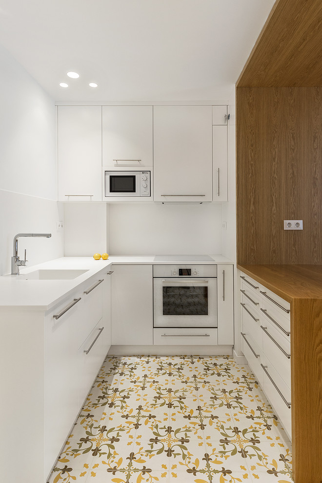 This is an example of a small contemporary u-shaped kitchen in Barcelona with an integrated sink, flat-panel cabinets, white cabinets, white splashback, white appliances, ceramic flooring, a breakfast bar, multi-coloured floors and white worktops.