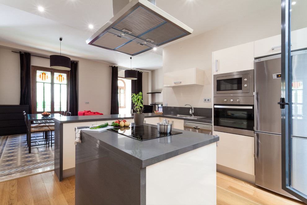 Photo of a medium sized contemporary l-shaped kitchen in Seville with a built-in sink, flat-panel cabinets, white cabinets, stainless steel appliances, light hardwood flooring, an island, composite countertops and grey splashback.