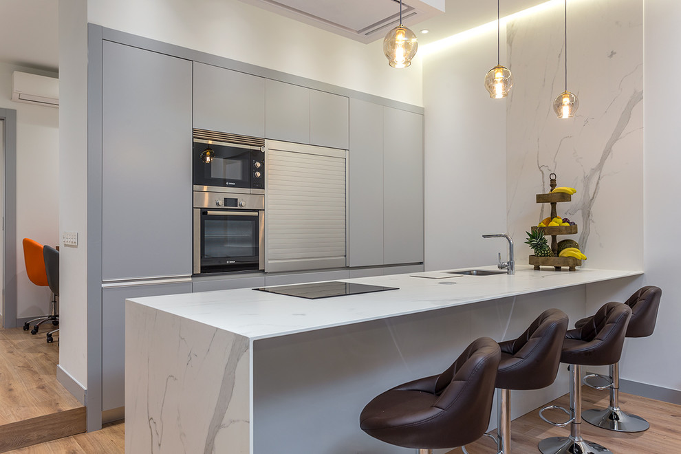 Photo of a medium sized contemporary single-wall kitchen in Madrid with flat-panel cabinets, grey cabinets, marble worktops, medium hardwood flooring, a breakfast bar, white worktops, a submerged sink and stainless steel appliances.