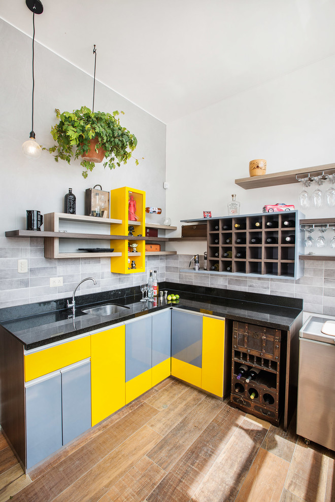 Пример оригинального дизайна: угловая кухня в современном стиле с врезной мойкой, плоскими фасадами, желтыми фасадами, серым фартуком, паркетным полом среднего тона, коричневым полом и черной столешницей
