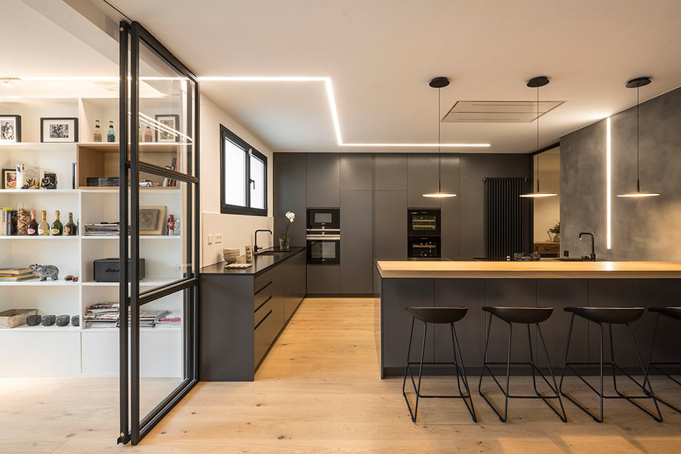 Идея дизайна: большая угловая кухня-гостиная в современном стиле с плоскими фасадами, черными фасадами, полуостровом, врезной мойкой, белым фартуком, черной техникой и светлым паркетным полом