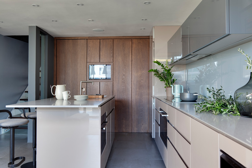 Contemporary kitchen in London with flat-panel cabinets, beige cabinets, white splashback, glass sheet splashback, black appliances, an island, grey floors and white worktops.