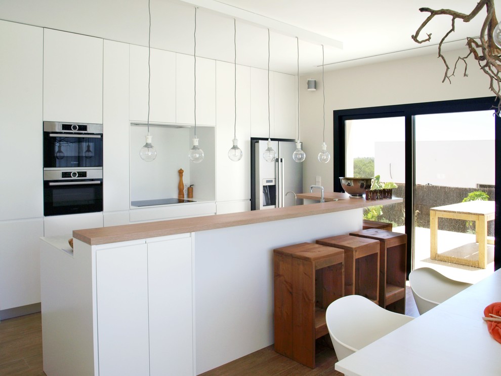 Modelo de cocina comedor minimalista con electrodomésticos de acero inoxidable, suelo de madera en tonos medios, encimeras blancas, una isla, armarios con paneles lisos, puertas de armario blancas y salpicadero blanco