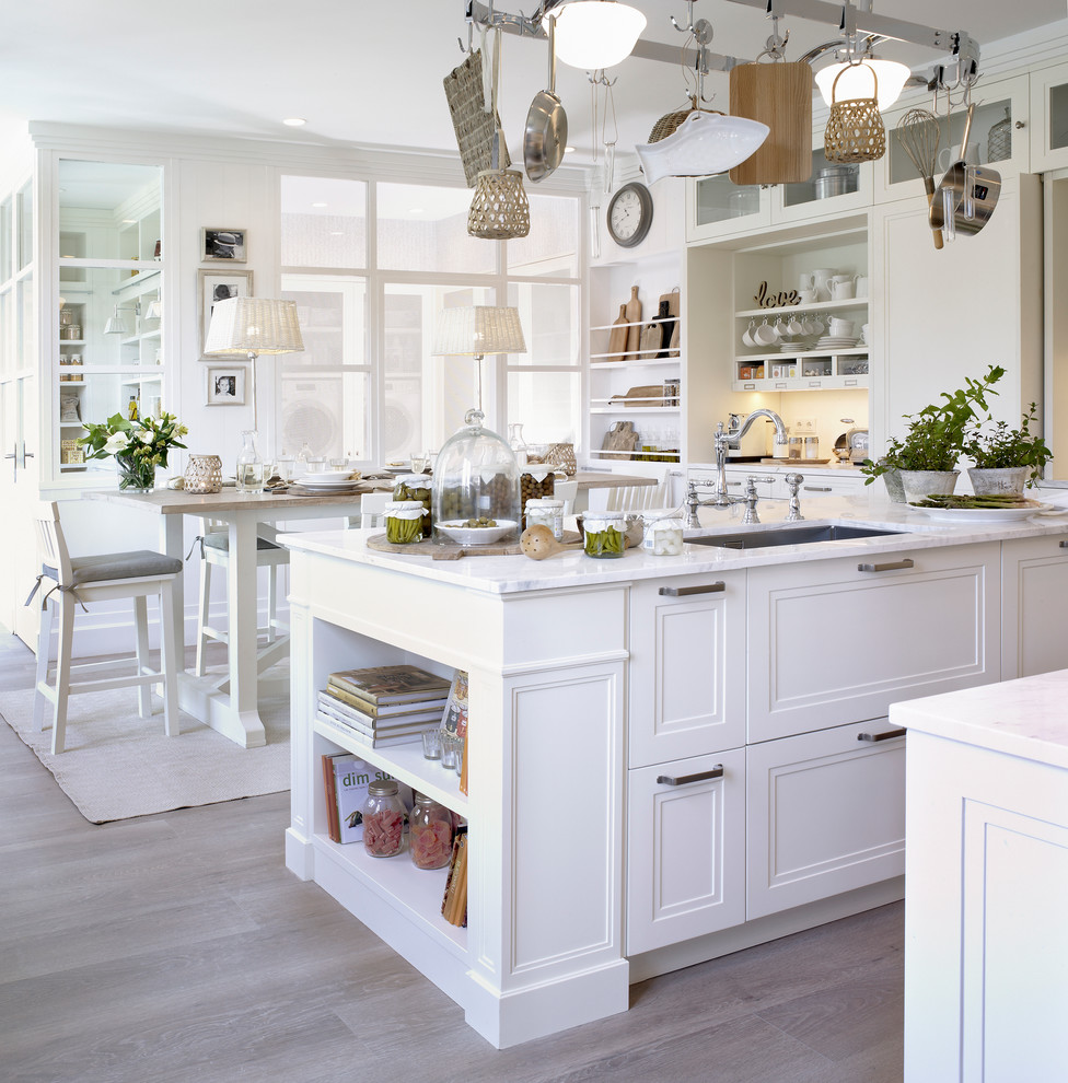 Lantlig inredning av ett stort kök, med en undermonterad diskho, vita skåp, marmorbänkskiva, vitt stänkskydd, stänkskydd i sten, vita vitvaror, ljust trägolv, en köksö och luckor med infälld panel