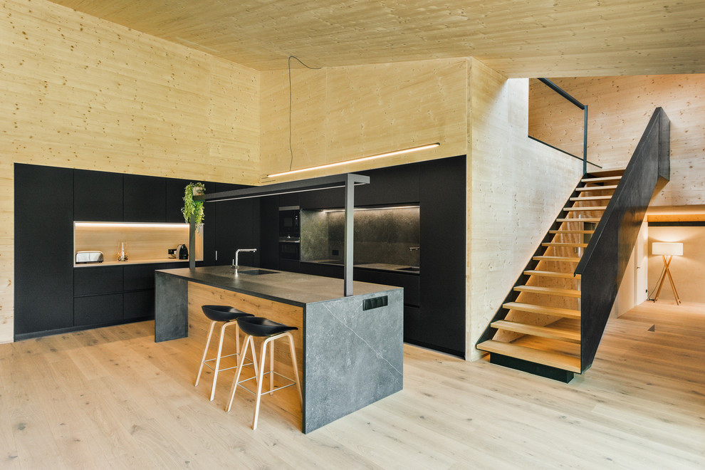 Идея дизайна: угловая кухня-гостиная в стиле модернизм с монолитной мойкой, плоскими фасадами, черными фасадами, черным фартуком, черной техникой, светлым паркетным полом, островом и черной столешницей
