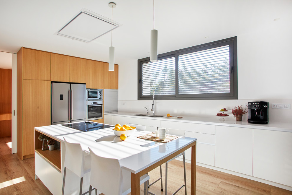 This is an example of a modern kitchen in Barcelona with flat-panel cabinets, white cabinets, engineered stone countertops, white splashback, stainless steel appliances, medium hardwood flooring, an island and white worktops.