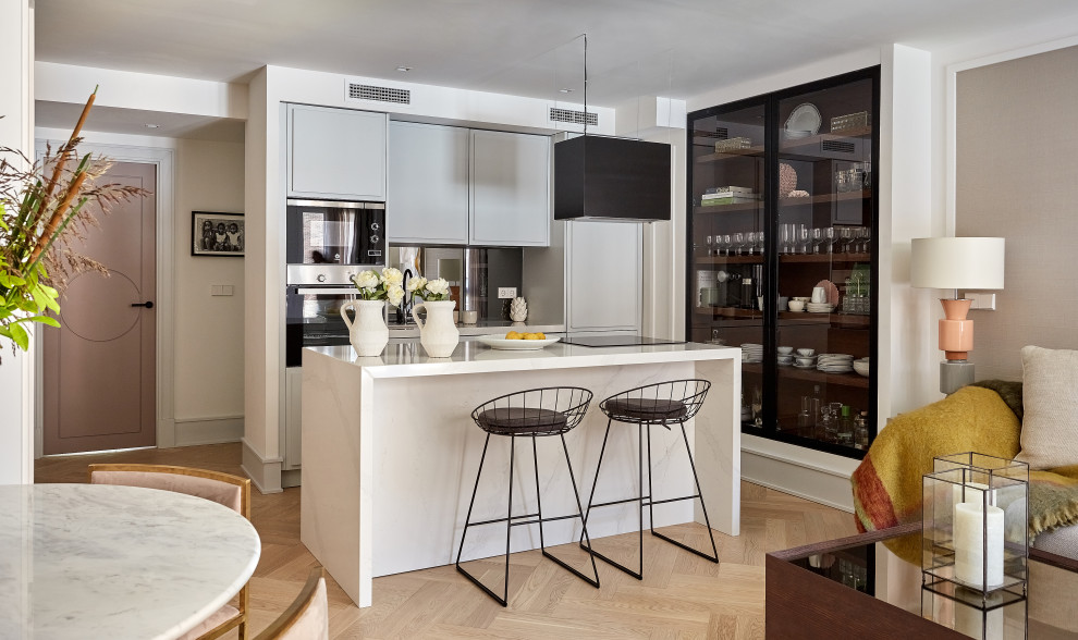 Cette image montre une petite cuisine ouverte parallèle design avec un placard à porte plane, des portes de placard grises, un électroménager en acier inoxydable, parquet clair, îlot, un sol beige et un plan de travail blanc.