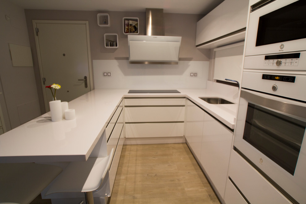 Design ideas for a small contemporary u-shaped open plan kitchen in Malaga with a single-bowl sink, flat-panel cabinets, white cabinets, white splashback, white appliances, medium hardwood flooring and no island.