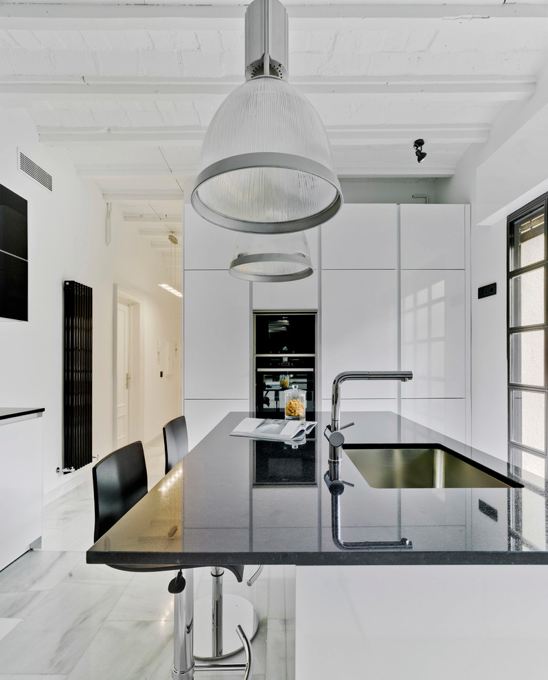 Bild på ett funkis kök, med en undermonterad diskho, släta luckor, vita skåp, granitbänkskiva, vitt stänkskydd, svarta vitvaror, en köksö, vitt golv och marmorgolv