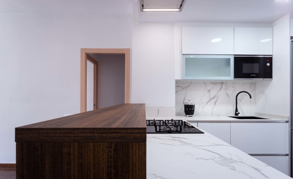 Design ideas for a small contemporary l-shaped open plan kitchen in Barcelona with a single-bowl sink, flat-panel cabinets, white cabinets, white splashback, stainless steel appliances, dark hardwood flooring, a breakfast bar, brown floors and white worktops.