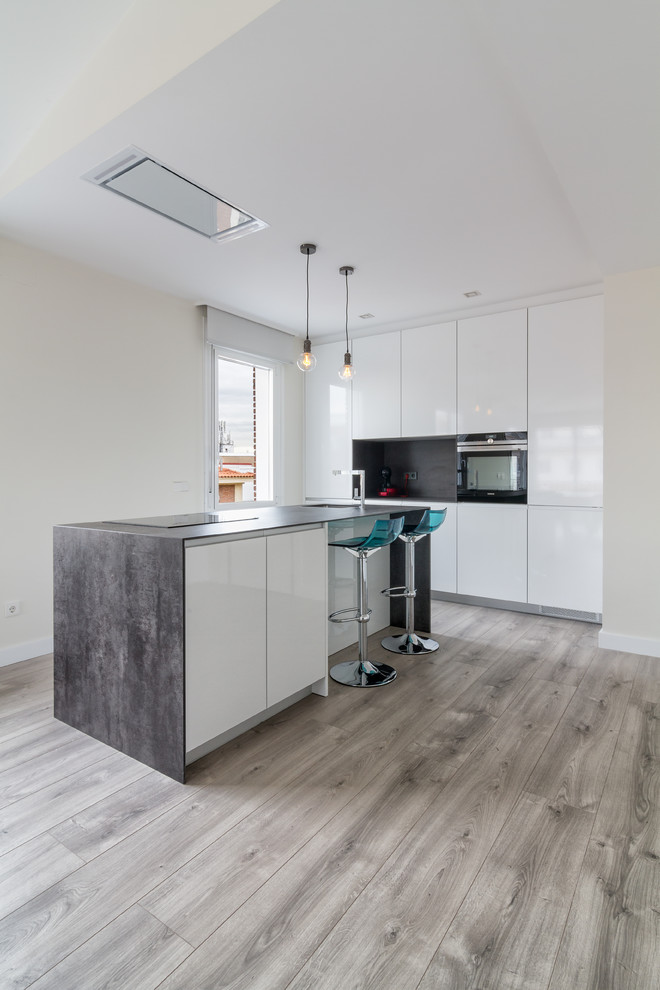 Contemporary single-wall open plan kitchen in Madrid with flat-panel cabinets, white cabinets, grey splashback, black appliances, medium hardwood flooring, an island, a submerged sink, grey floors and grey worktops.