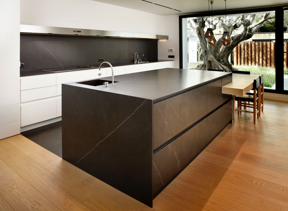 Inredning av ett modernt mycket stort svart linjärt svart kök med öppen planlösning, med en integrerad diskho, släta luckor, vita skåp, bänkskiva i kvarts, svart stänkskydd, rostfria vitvaror, ljust trägolv, en köksö och brunt golv