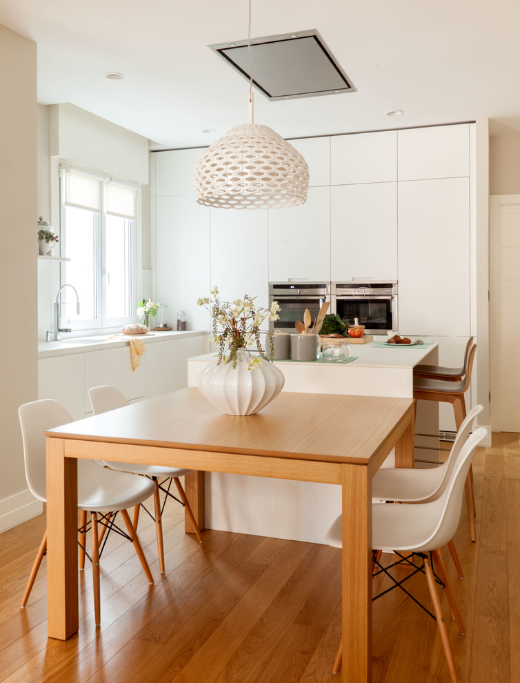 Mittelgroße Moderne Wohnküche in L-Form mit flächenbündigen Schrankfronten, weißen Schränken, Küchengeräten aus Edelstahl, Kücheninsel, braunem Boden, Küchenrückwand in Weiß und braunem Holzboden in Bilbao