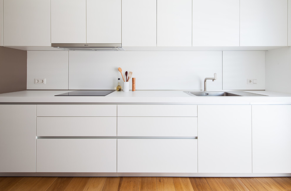 Bild på ett funkis kök, med en enkel diskho, släta luckor, vita skåp, laminatbänkskiva, vitt stänkskydd, rostfria vitvaror, mellanmörkt trägolv och brunt golv