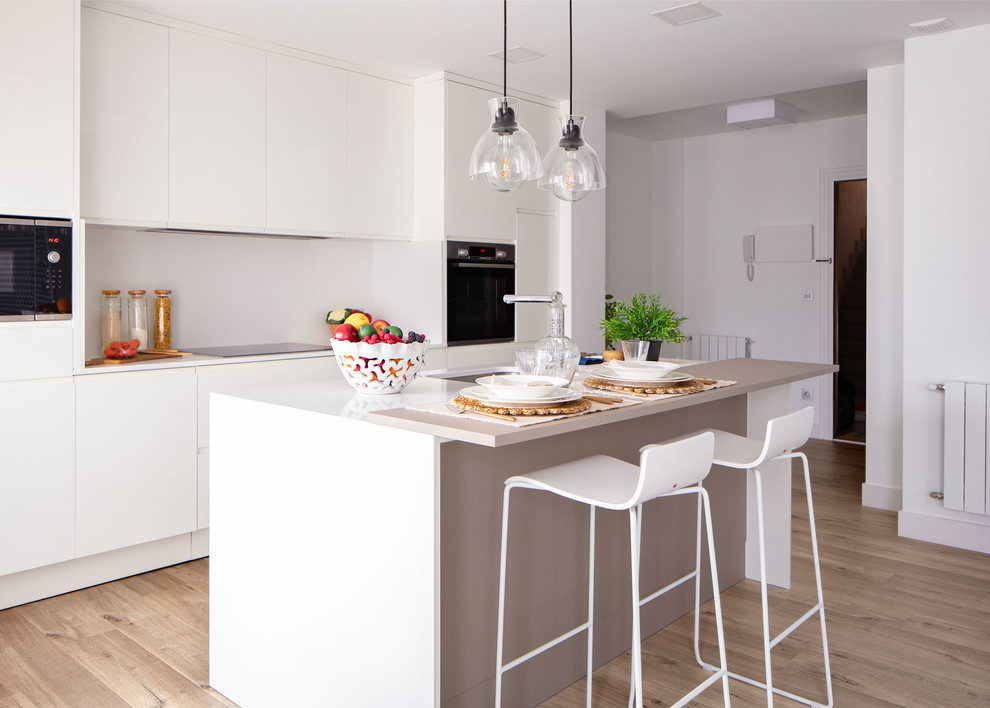 Bild på ett mellanstort funkis vit linjärt vitt kök med öppen planlösning, med en undermonterad diskho, släta luckor, vita skåp, marmorbänkskiva, vitt stänkskydd, stänkskydd i marmor, integrerade vitvaror, ljust trägolv, en köksö och brunt golv
