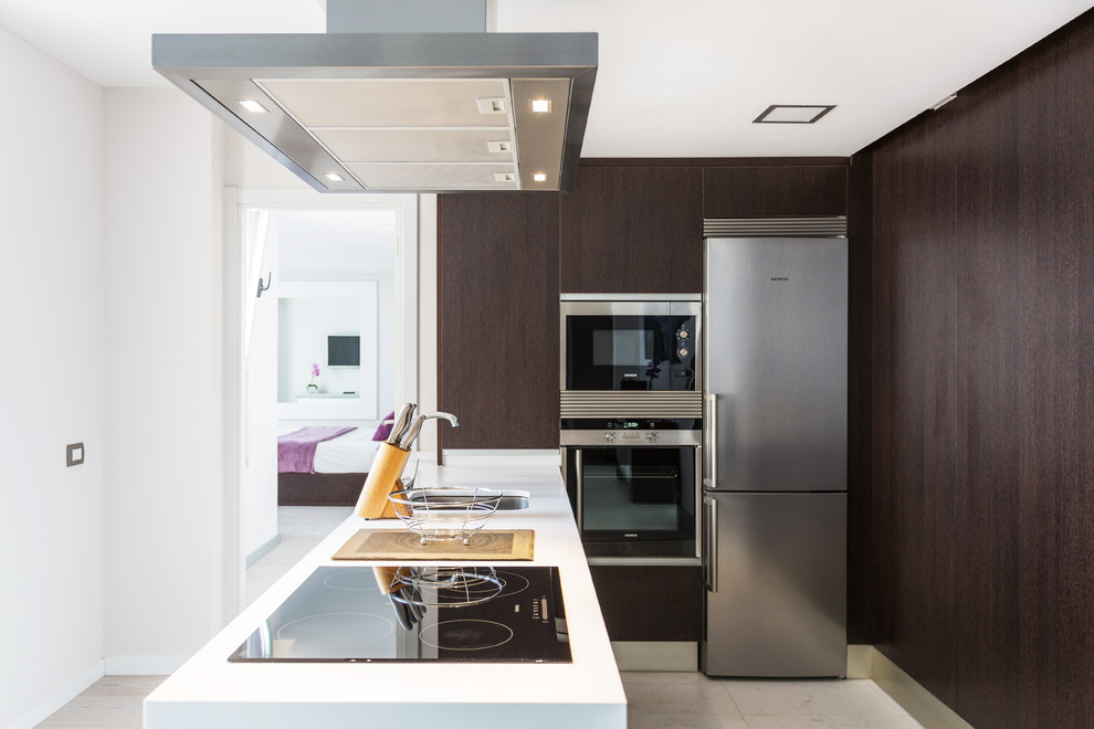 Small beach style l-shaped travertine floor enclosed kitchen photo in Other with an undermount sink, solid surface countertops, stainless steel appliances, a peninsula, flat-panel cabinets, dark wood cabinets and white countertops