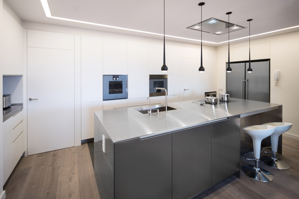 Example of a large trendy single-wall dark wood floor and brown floor eat-in kitchen design in Barcelona with an integrated sink, flat-panel cabinets, white cabinets, stainless steel countertops, gray backsplash, metal backsplash, stainless steel appliances, an island and gray countertops