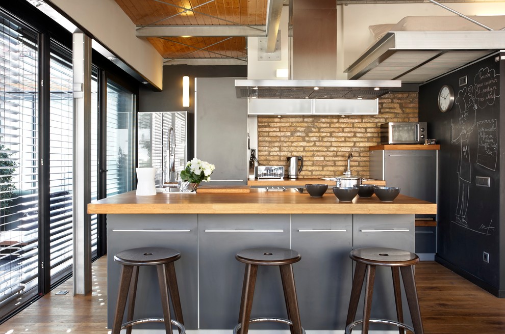 Exemple d'une cuisine ouverte industrielle avec un placard à porte plane, des portes de placard grises, un plan de travail en bois, une crédence en brique, un électroménager en acier inoxydable, un sol en bois brun et îlot.