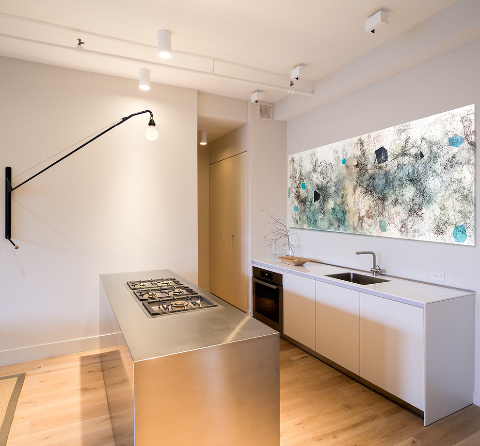 Inspiration for a small contemporary galley medium tone wood floor kitchen remodel in New York with an undermount sink, flat-panel cabinets, white cabinets, stainless steel countertops, stainless steel appliances and an island