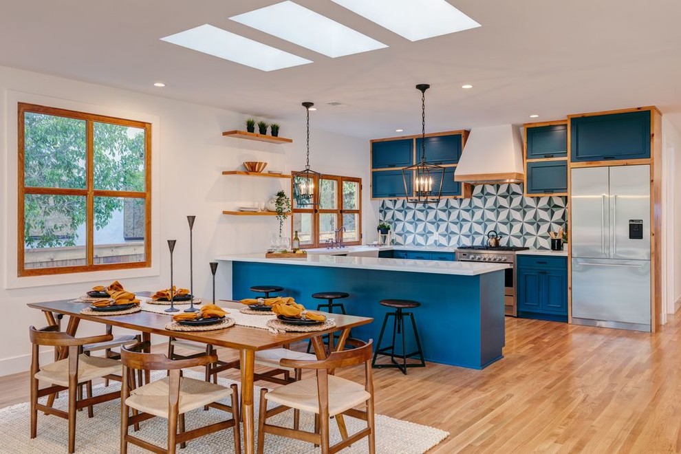 Large world-inspired single-wall open plan kitchen in Los Angeles with a double-bowl sink, recessed-panel cabinets, medium wood cabinets, granite worktops, multi-coloured splashback, porcelain splashback, stainless steel appliances, medium hardwood flooring, an island, brown floors and white worktops.