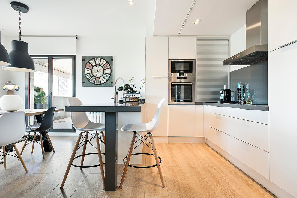 Mittelgroße Moderne Wohnküche in U-Form mit flächenbündigen Schrankfronten, weißen Schränken, Küchenrückwand in Grau, Küchengeräten aus Edelstahl, hellem Holzboden und Halbinsel in Barcelona