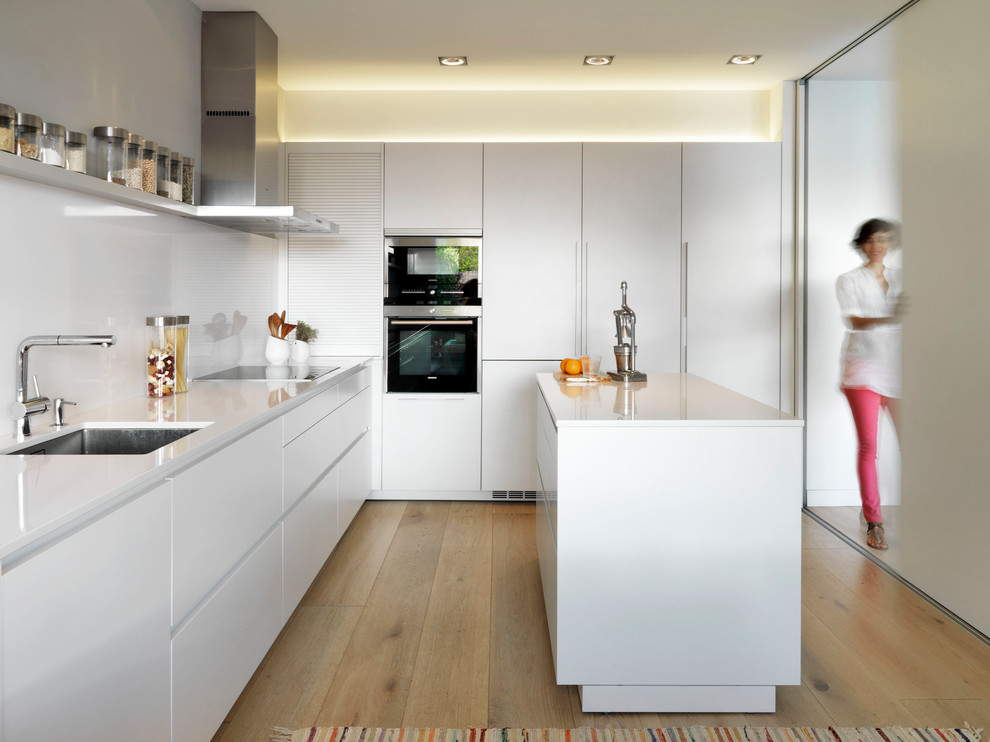 Photo of a medium sized contemporary single-wall kitchen/diner in Madrid with a submerged sink, flat-panel cabinets, white cabinets, white splashback, stainless steel appliances, an island and light hardwood flooring.