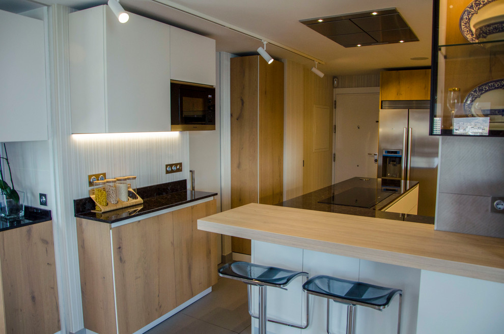Photo of a medium sized u-shaped kitchen/diner in Malaga with a belfast sink, glass-front cabinets, medium wood cabinets, granite worktops, stainless steel appliances, ceramic flooring, a breakfast bar, beige floors and black worktops.