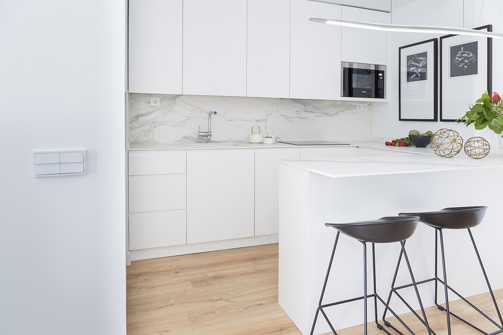 Contemporary u-shaped kitchen in Madrid with a submerged sink, flat-panel cabinets, white cabinets, light hardwood flooring, a breakfast bar, white worktops and beige floors.