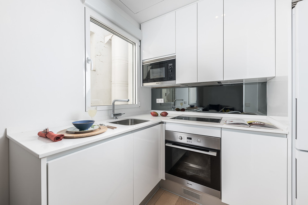 Inredning av ett minimalistiskt litet vit vitt kök, med en undermonterad diskho, släta luckor, vita skåp, bänkskiva i kvarts, grått stänkskydd, fönster som stänkskydd, rostfria vitvaror, ljust trägolv och beiget golv