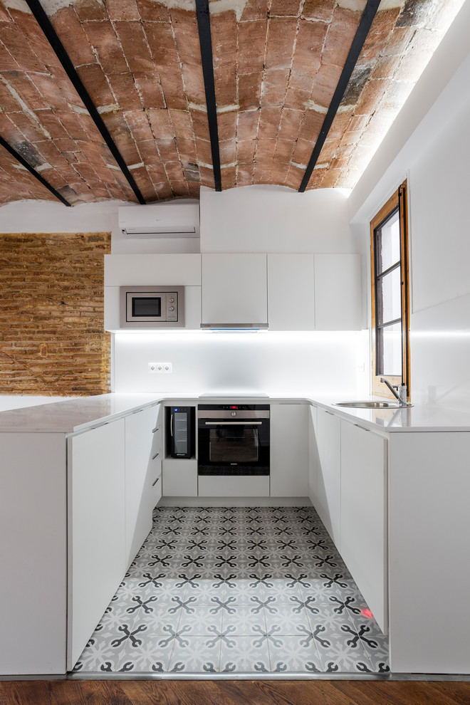 バルセロナにある小さな地中海スタイルのおしゃれなキッチン (フラットパネル扉のキャビネット、白いキャビネット、白いキッチンパネル、シルバーの調理設備、セラミックタイルの床、ドロップインシンク、グレーの床、白いキッチンカウンター) の写真