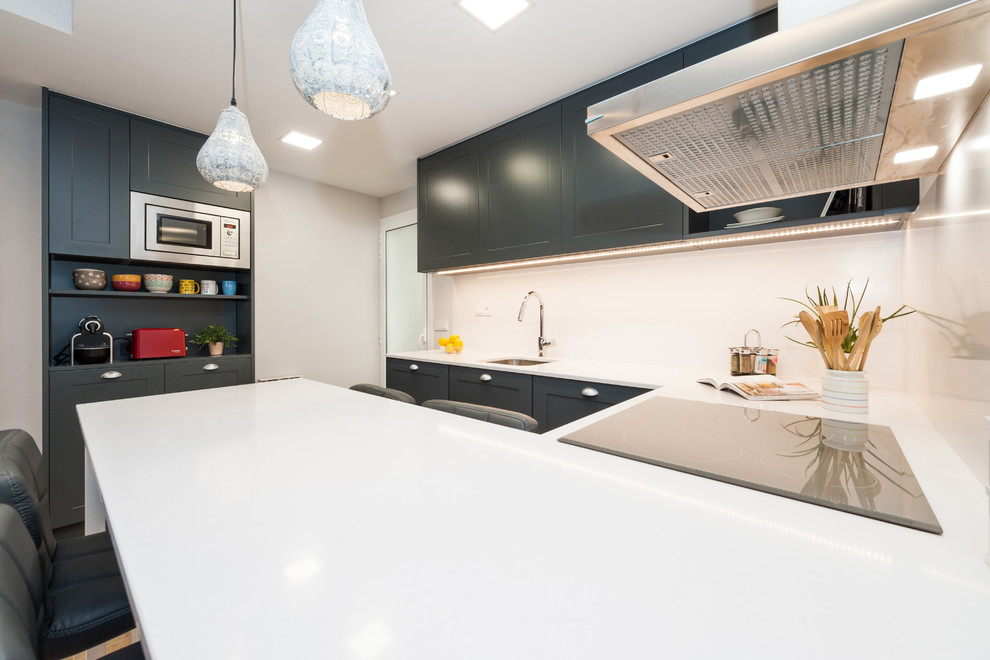 Photo of a medium sized traditional u-shaped open plan kitchen in Barcelona with a single-bowl sink, recessed-panel cabinets, blue cabinets, engineered stone countertops, white splashback, stainless steel appliances, porcelain flooring, a breakfast bar, brown floors and white worktops.
